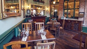 Un restaurante con mesas y sillas de madera, una zona de bar y una pared de ladrillo con un gran ventanal arqueado.