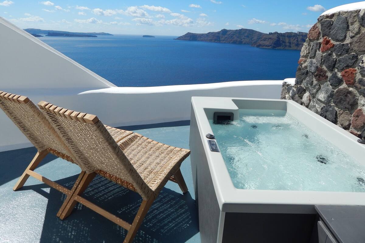 A sunlit outdoor area with a wicker chair and a hot tub overlooking a body of water and distant landforms.