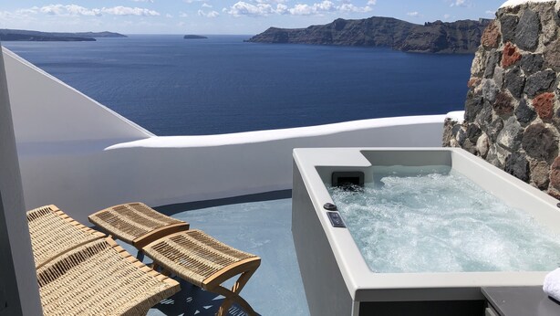A hot tub with a stone wall backdrop, overlooking a body of water and distant land. - Aspa Caves (Santorini)