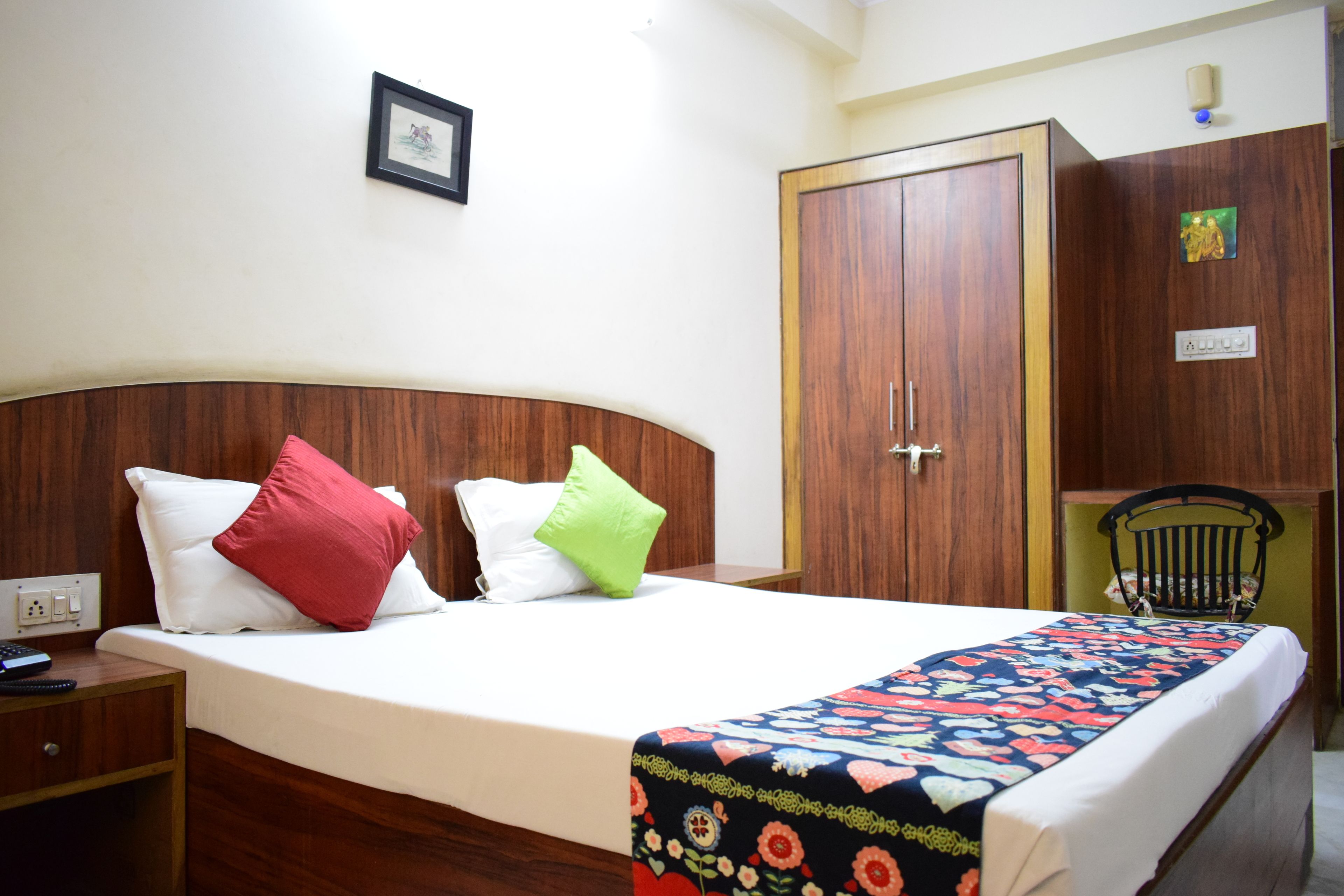 A hotel room with a wooden headboard, a bed with white bedding, a floral-patterned bedspread, a nightstand, a wardrobe, and a chair.