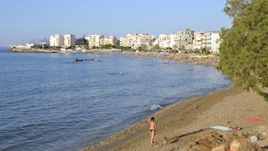A beach with a person walking along the shore, buildings in the background, and a clear sky. - Elia Seaside Nea Chora (Chania)