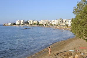 Μια παραλία με ένα άτομο να περπατά κατά μήκος της ακτής, κτίρια στο βάθος και καθαρός ουρανός.