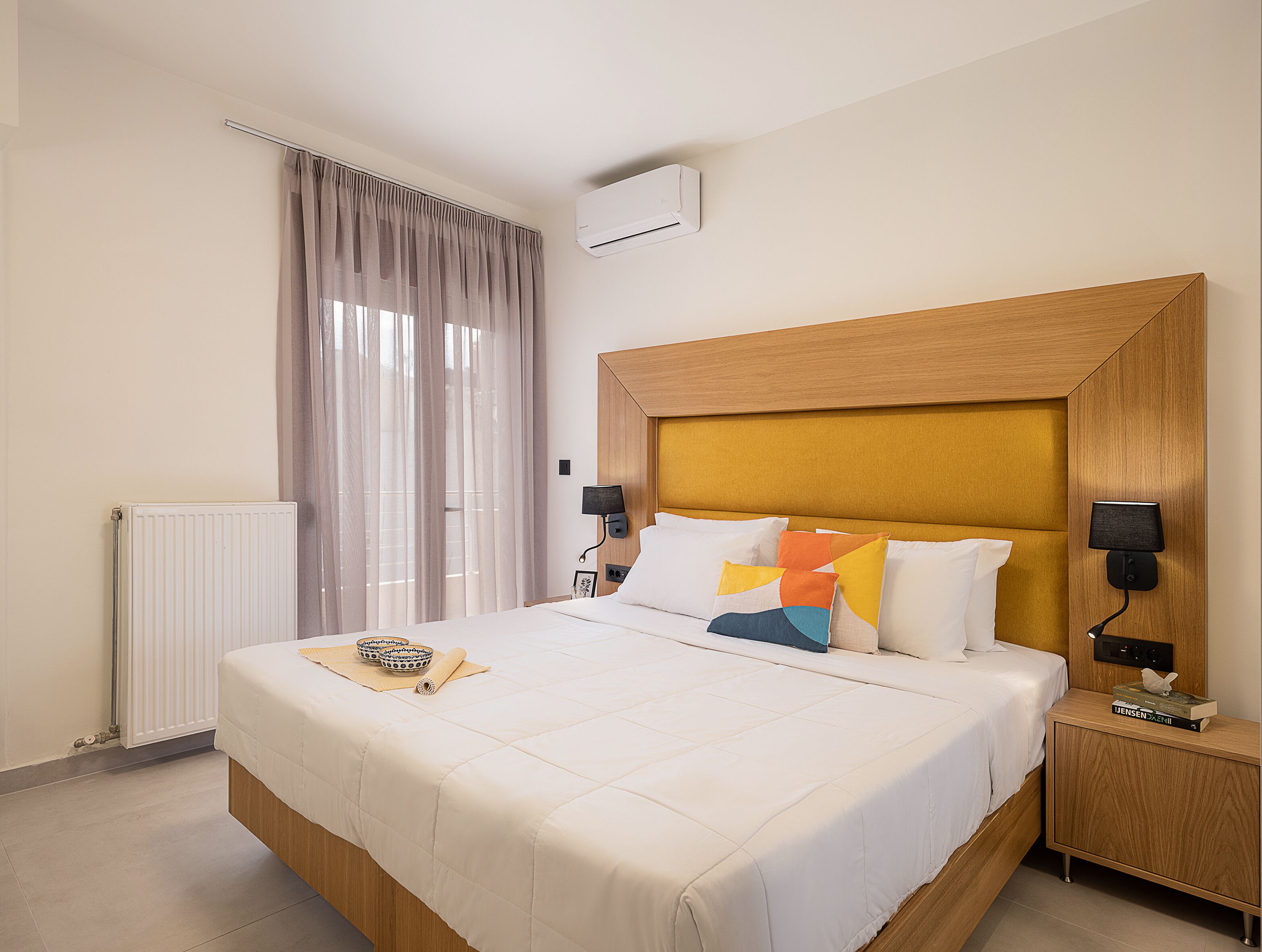 A hotel room with a large bed, a wooden headboard, two bedside lamps, a radiator, and a window with curtains.