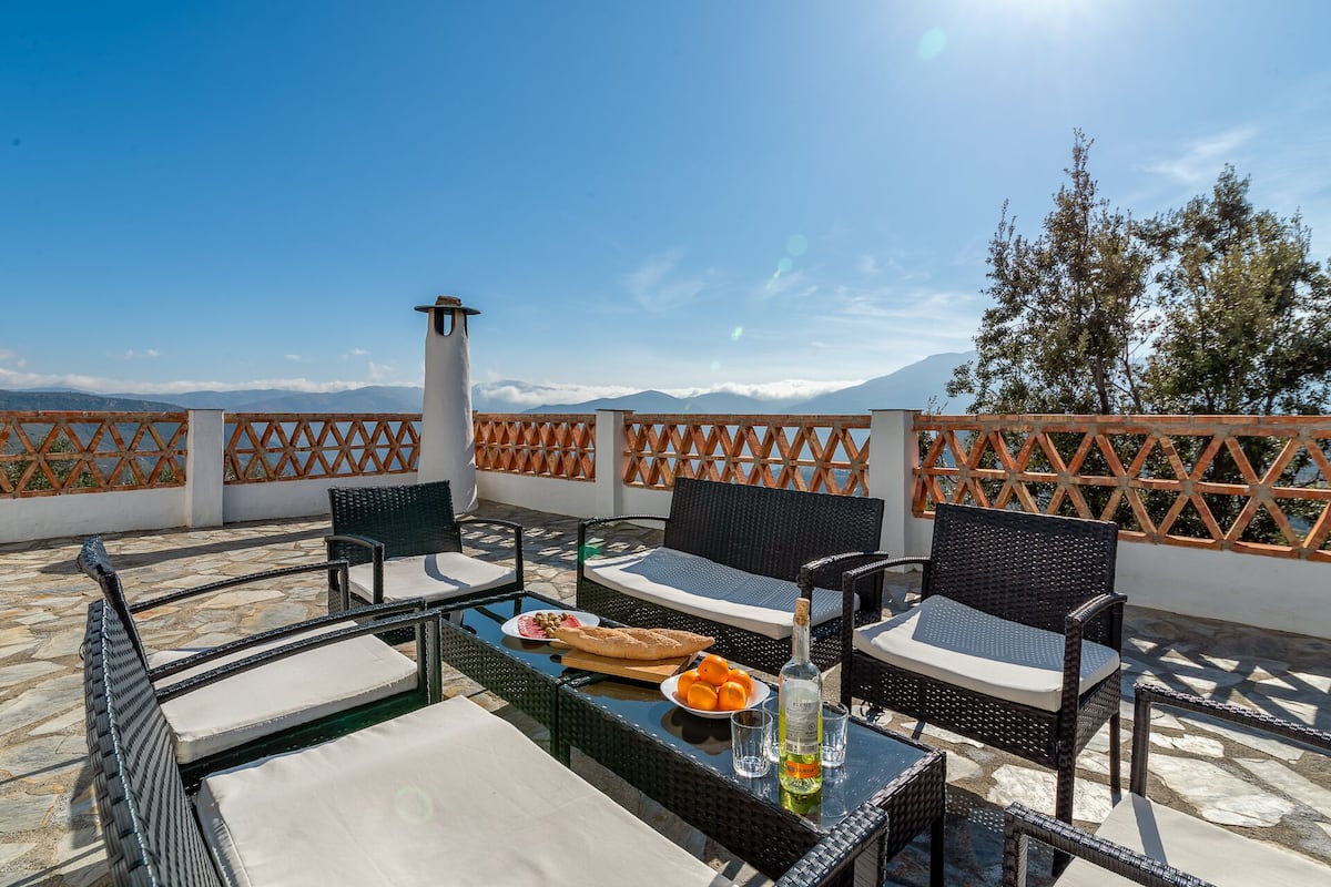 Une terrasse avec des meubles en osier, une bouteille de vin et une assiette d’oranges.