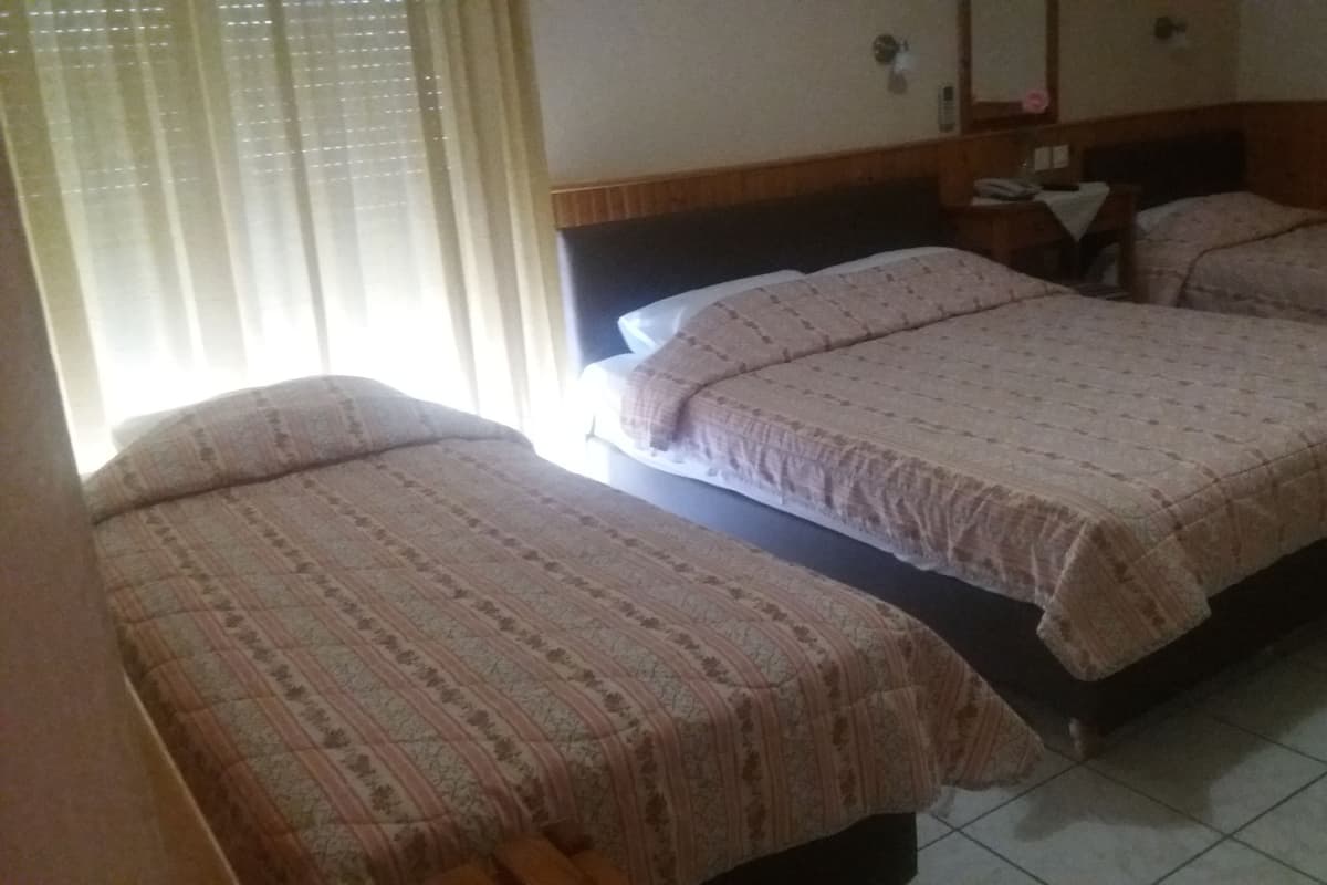 Habitación de hotel con tres camas individuales, cabecera de madera, mesita de noche con lámpara y ventana con persianas.