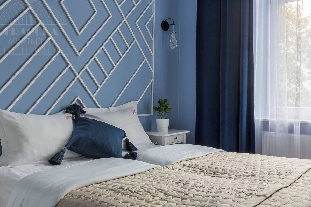 Un dormitorio con un cabecero de estampado geométrico, una mesita de noche con una planta y una ventana con cortinas translúcidas.