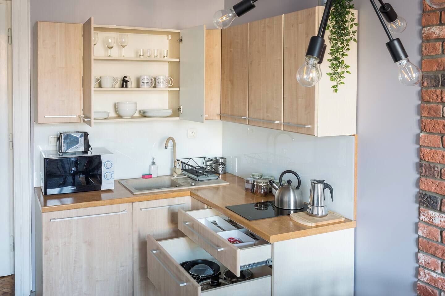 Cocina compacta con armarios de madera, un tostador integrado, un lavabo y una tetera.