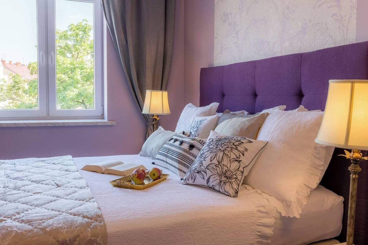 Una cama bien hecha con almohadas y una bandeja de manzanas, una ventana con cortinas y dos lámparas de noche.