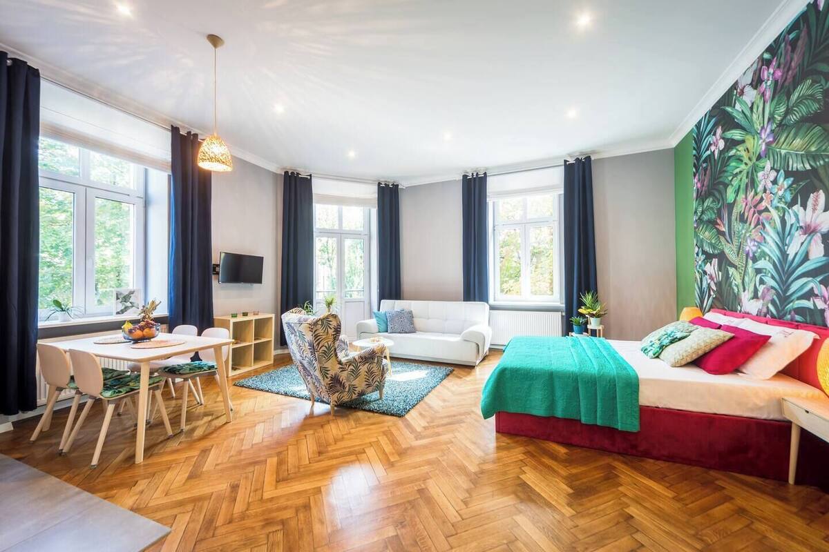 Amplia habitación con suelo de madera en patrón de espiga, cama con colcha verde, sofá blanco, mesa de comedor con sillas y un ventanal con cortinas.
