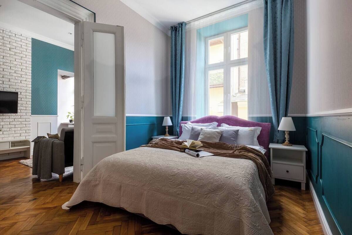 Un dormitorio con cama, mesitas de noche, ventana con cortinas, puerta y pared de ladrillo.