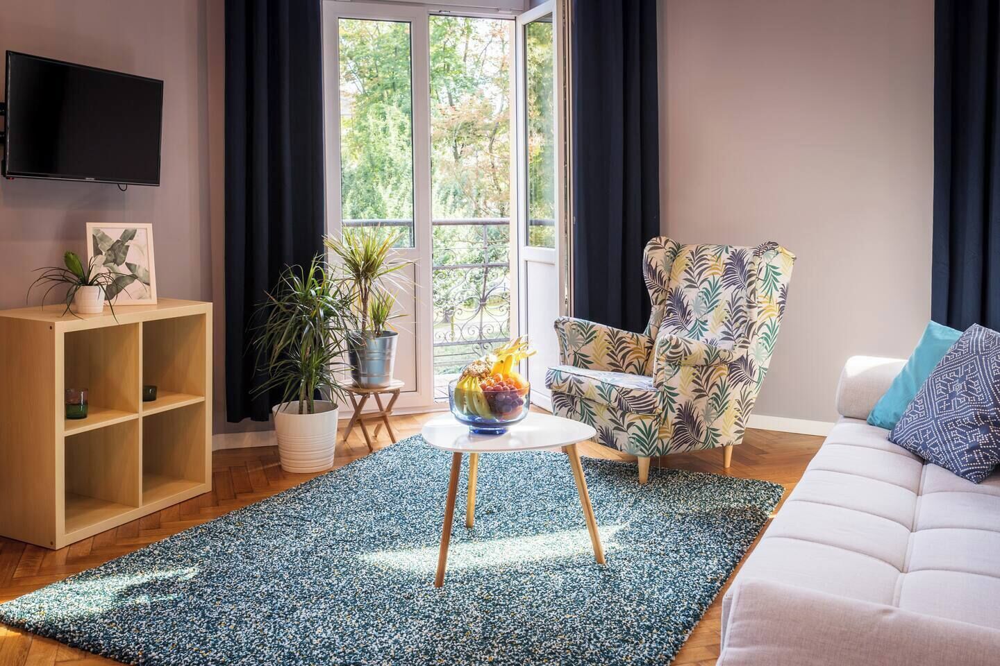 Una sala de estar con un sofá, un sillón estampado, una mesa de centro con fruta, un televisor y una ventana con cortinas.