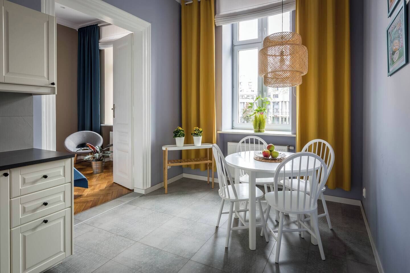 A modern kitchen with white cabinets, a dining table with chairs, a hanging light fixture, and a window with yellow curtains.