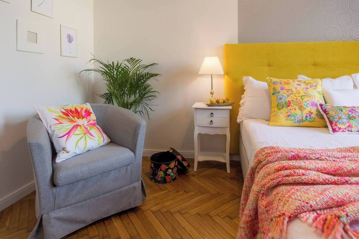 A room with a grey armchair, a bed with a yellow headboard, a small bedside table with a lamp, and a potted plant.