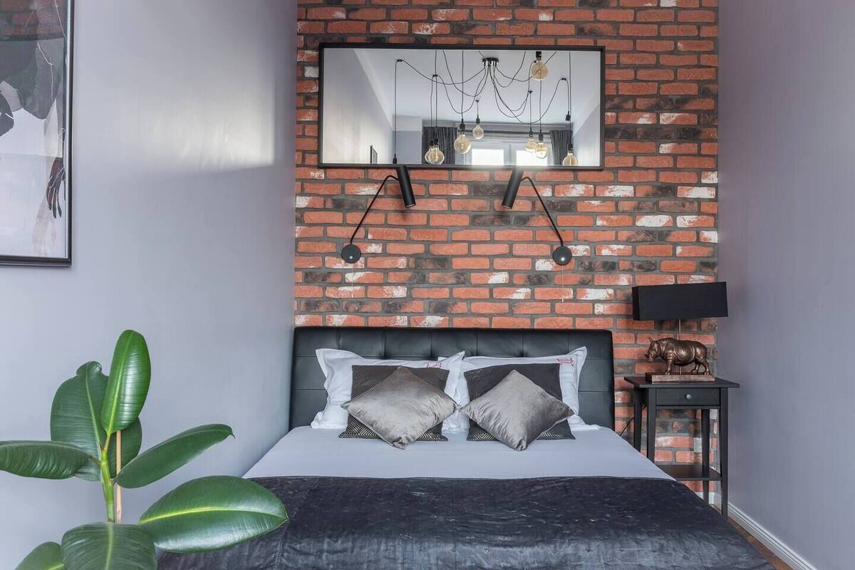 Un dormitorio con pared de ladrillo, cama con muchos almohadones, mesita de noche con lámpara y un espejo grande.