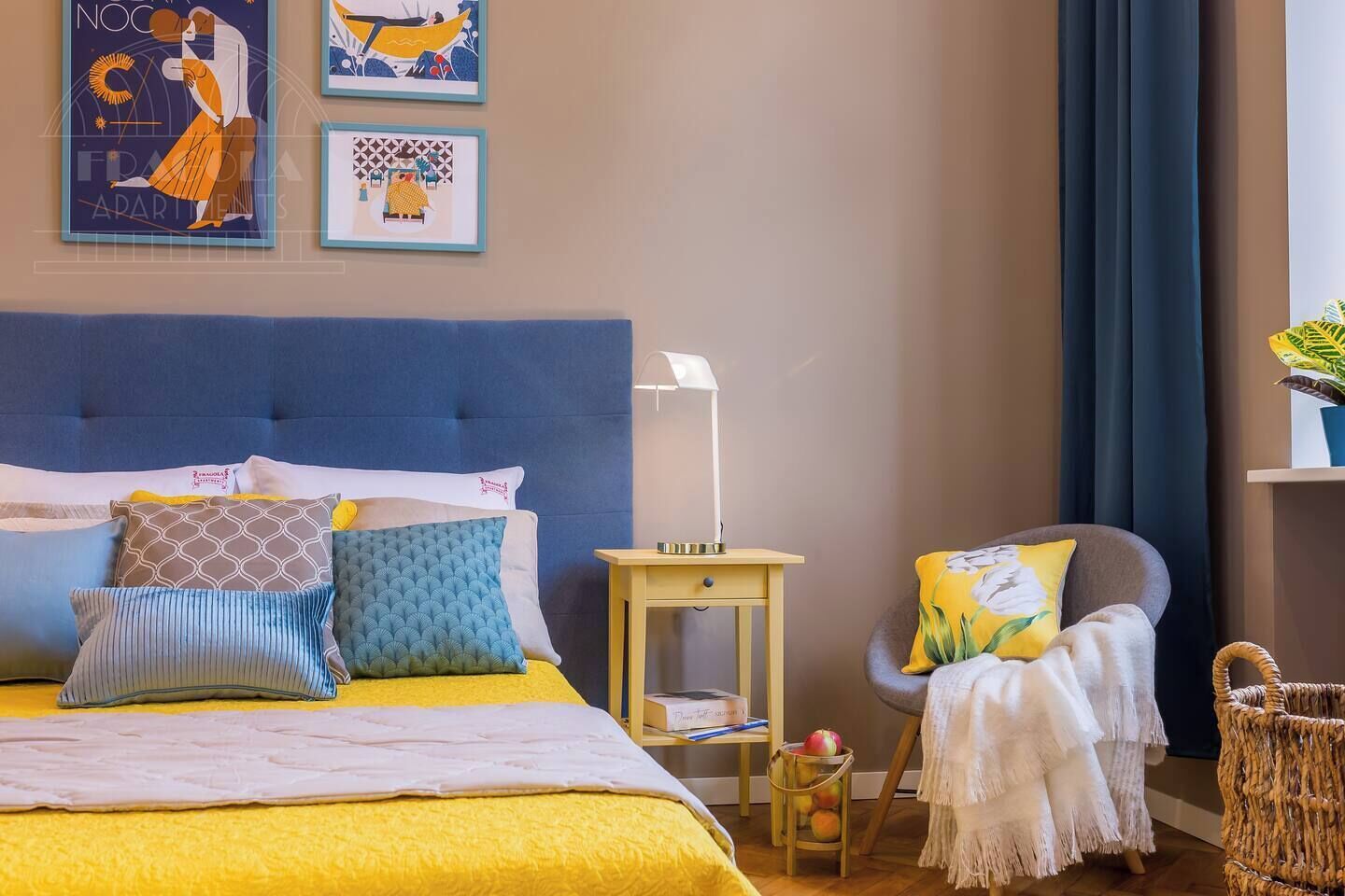 A bedroom with a bed, pillows, a bedside table, a lamp, a chair, and a wall with artwork.