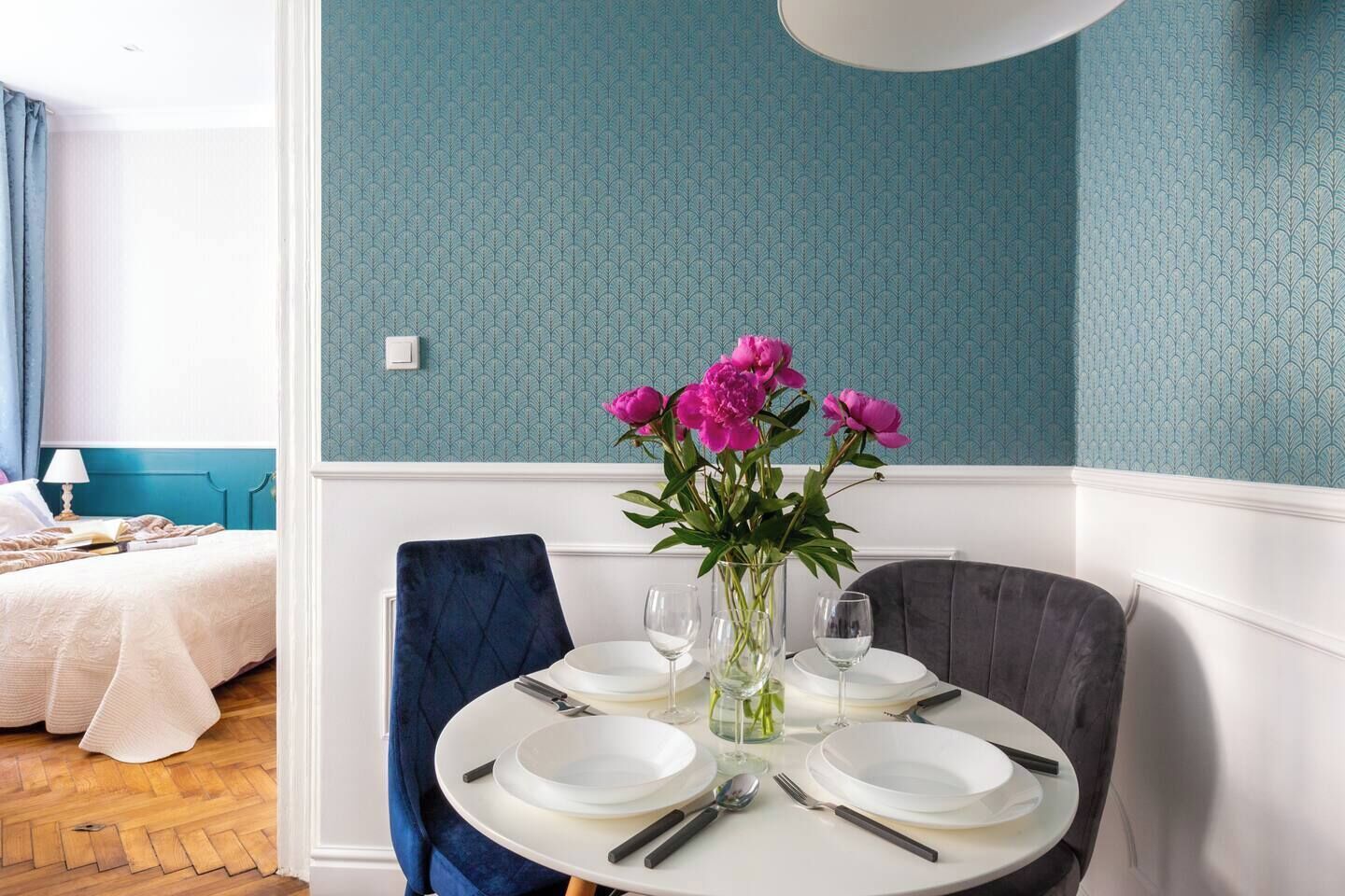 A dining area with a round table set for four, a vase of flowers, and a bed with a blue headboard in the background.