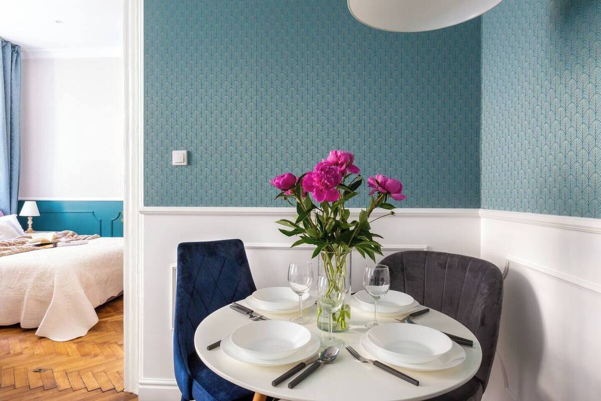 A dining area with a round table set for four, a vase of flowers, and a bed with a blue headboard in the background.