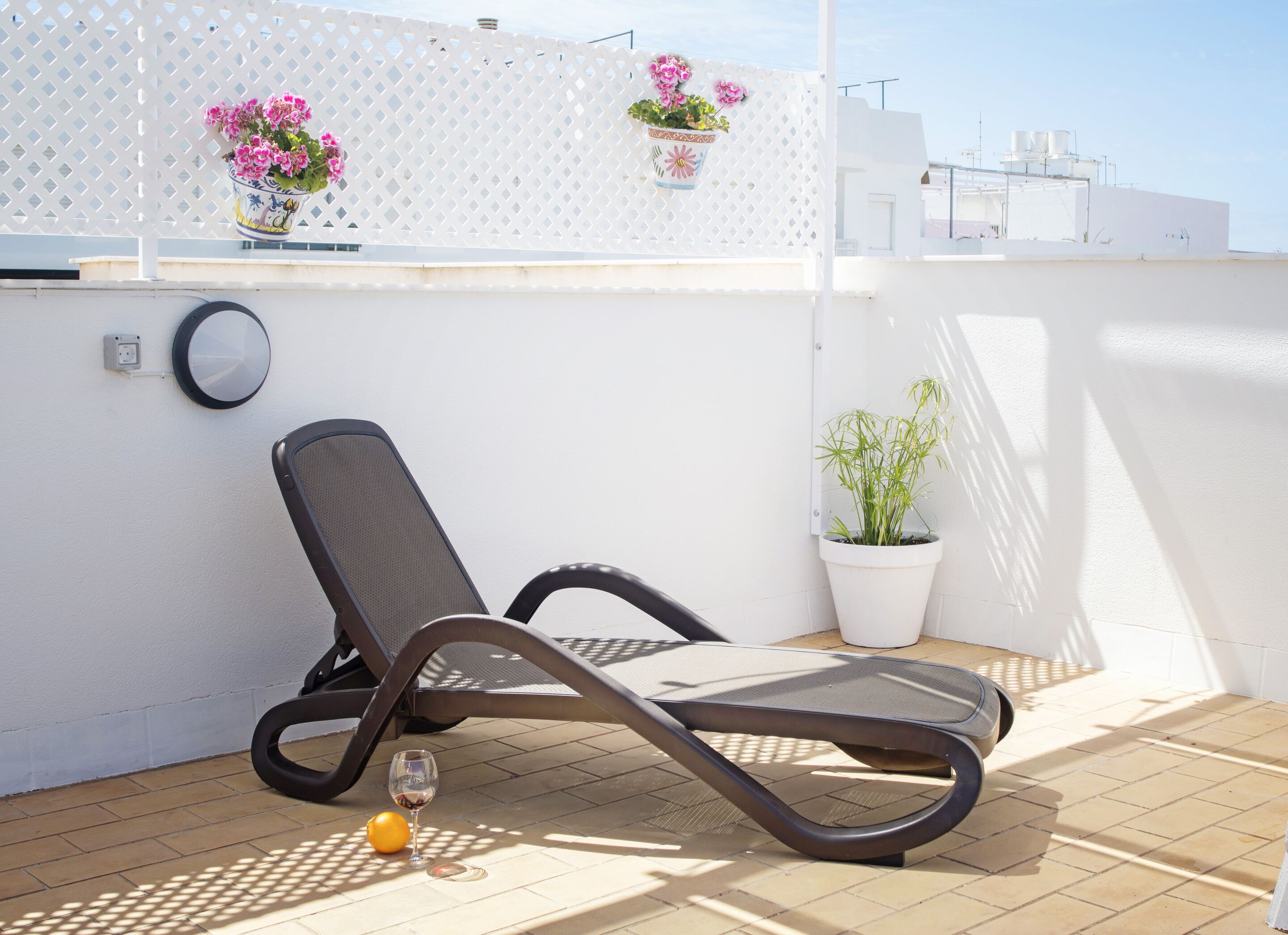 A sunlit outdoor seating area with a lounge chair, a potted plant, and a wine glass on the floor.