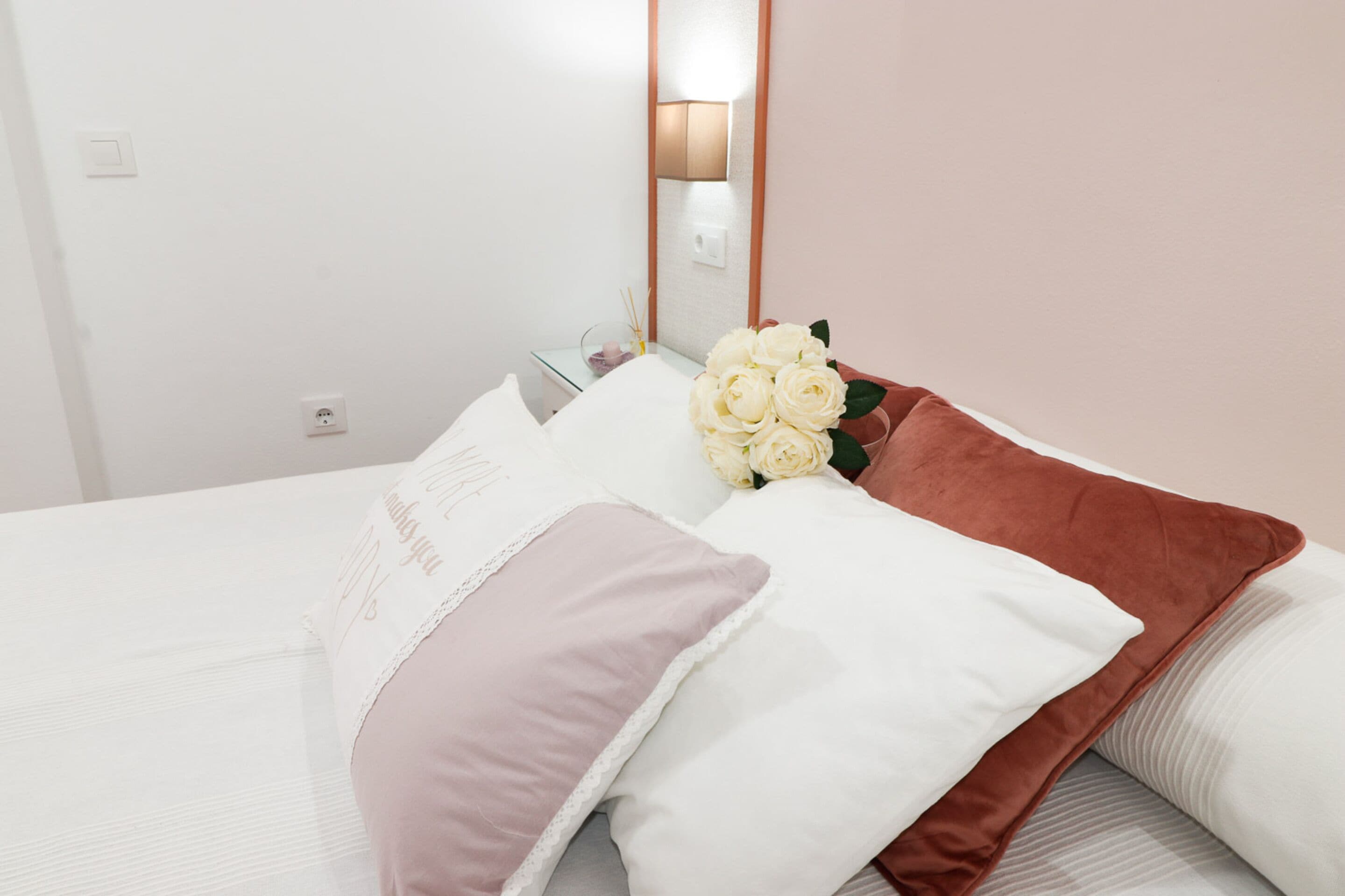 Een net opgemaakt bed met witte en roze kussens, een bruine plaid en een boeket witte rozen.