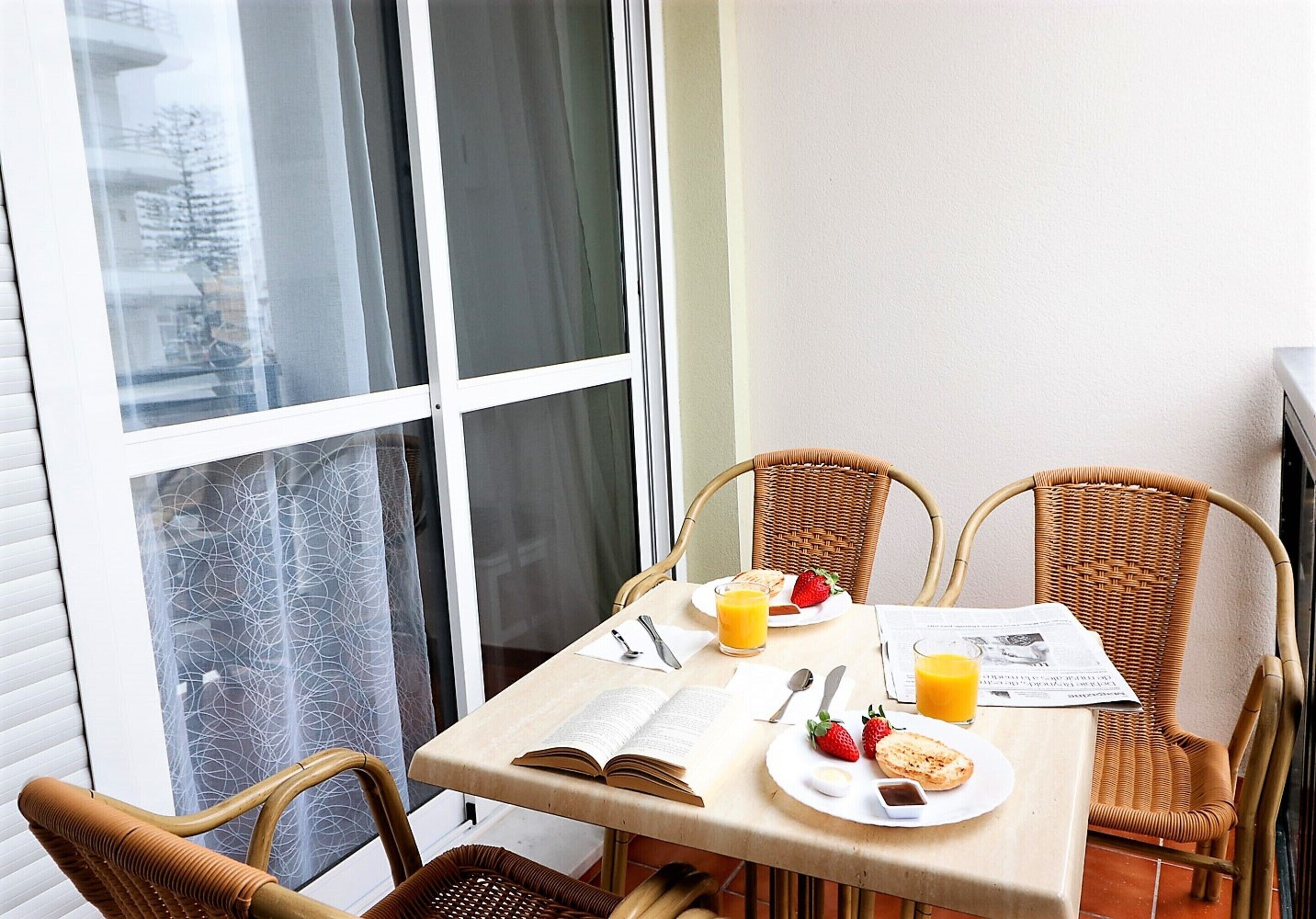 Een eettafel met rieten stoelen, een boek, een bord met eten en een glas sinaasappelsap, en een krant.