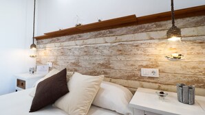 Une chambre à coucher avec une tête de lit en bois rustique, un lit recouvert de linge de maison blanc, deux tables de chevet ornées d’objets décoratifs et des lampes suspendues.