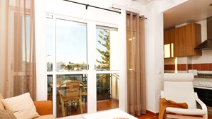 Un salon comprenant un canapé, une table basse et un fauteuil. Il y a un balcon avec une porte vitrée, une table à manger avec des chaises, et un coin cuisine avec des placards et une cuisinière.