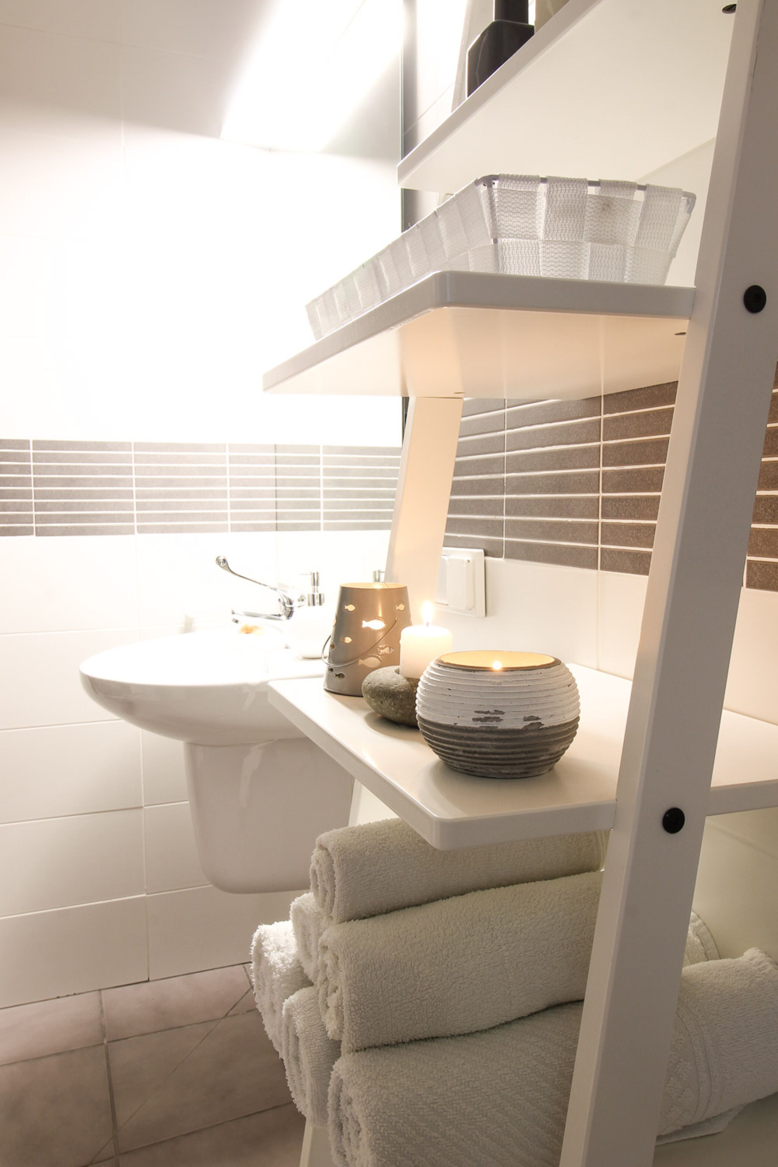 A bathroom with a white sink, a mirror, a shelf with towels, a candle, and a white bathtub.