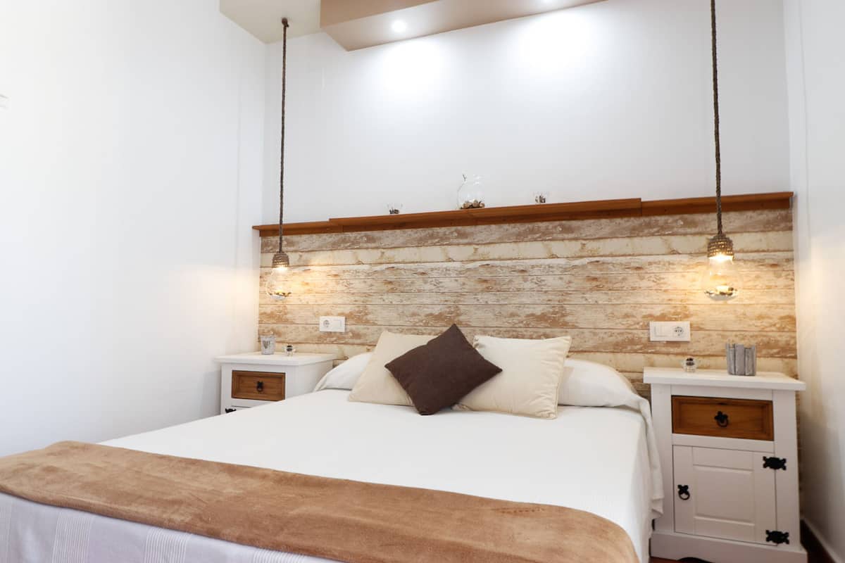 Een slaapkamer met een houten hoofdbord, twee nachtkastjes met lades, een bed met wit beddengoed en een bruine plaid, en twee hanglampen.