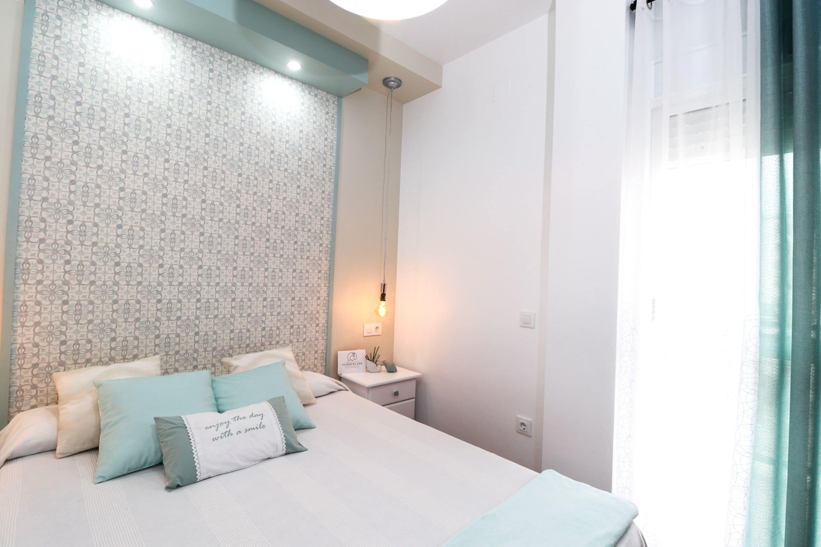 A neatly arranged bedroom with a bed, pillows, a bedside table with a lamp, and a patterned wall.