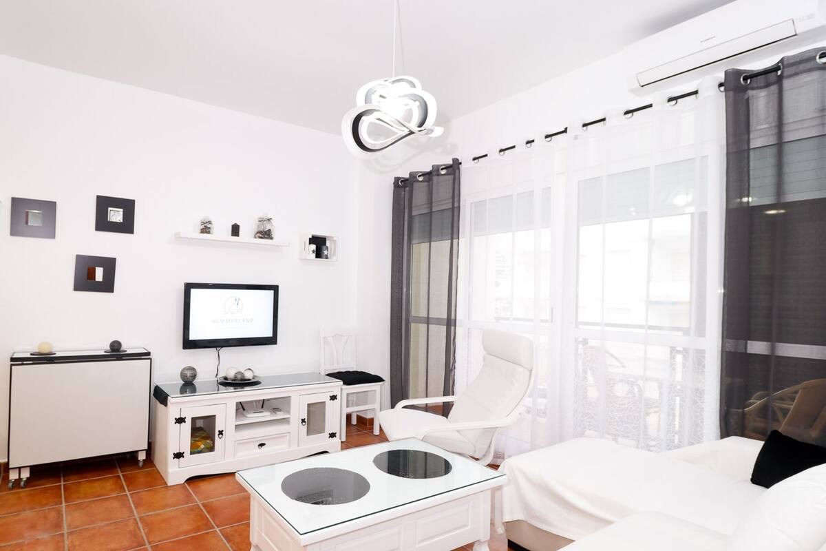 Een moderne, goed verlichte woonkamer met een witte bank, een glazen salontafel, een witte televisie en een eethoek met een wit kastje en een kleine tafel.