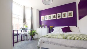A bedroom with a white bed, purple walls, and a window with curtains. There are framed pictures on the wall and a small table with a plant. - Modern Design House (Liverpool)