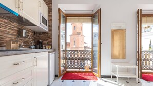 Una cocina moderna con armarios blancos, un microondas y una alfombra roja que lleva a un balcón con vistas a una torre de iglesia.