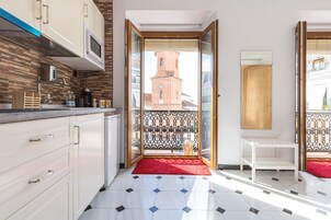 Una cocina moderna con armarios blancos, un microondas y una alfombra roja que lleva a un balcón con vistas a una torre de iglesia.