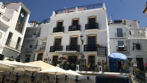 Un edificio de varios pisos con balcones, un reloj y zonas de estar al aire libre con sombrillas.