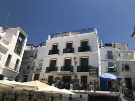 Un edificio de varios pisos con balcones, un reloj y zonas de estar al aire libre con sombrillas.