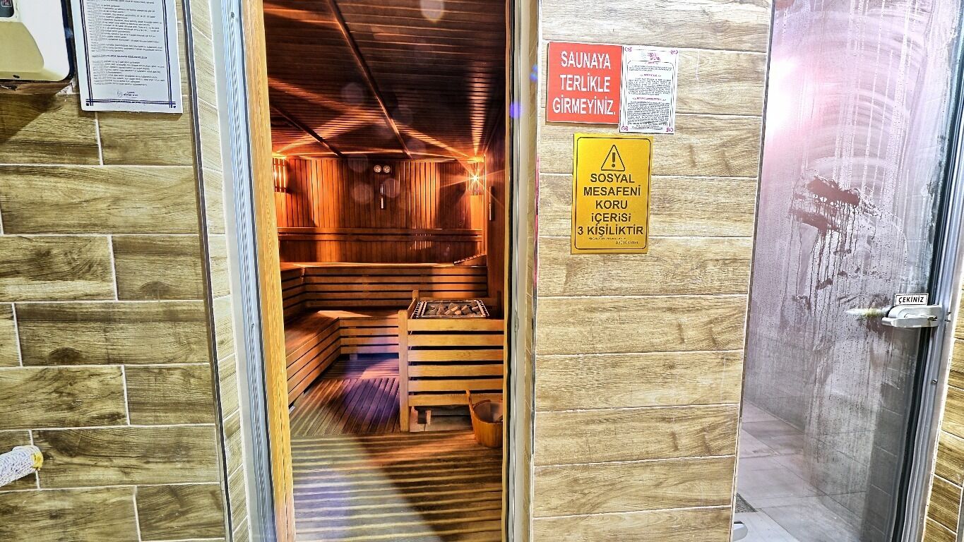 A sauna room with wooden walls and benches, a fire pit, and a sign indicating the sauna is open.