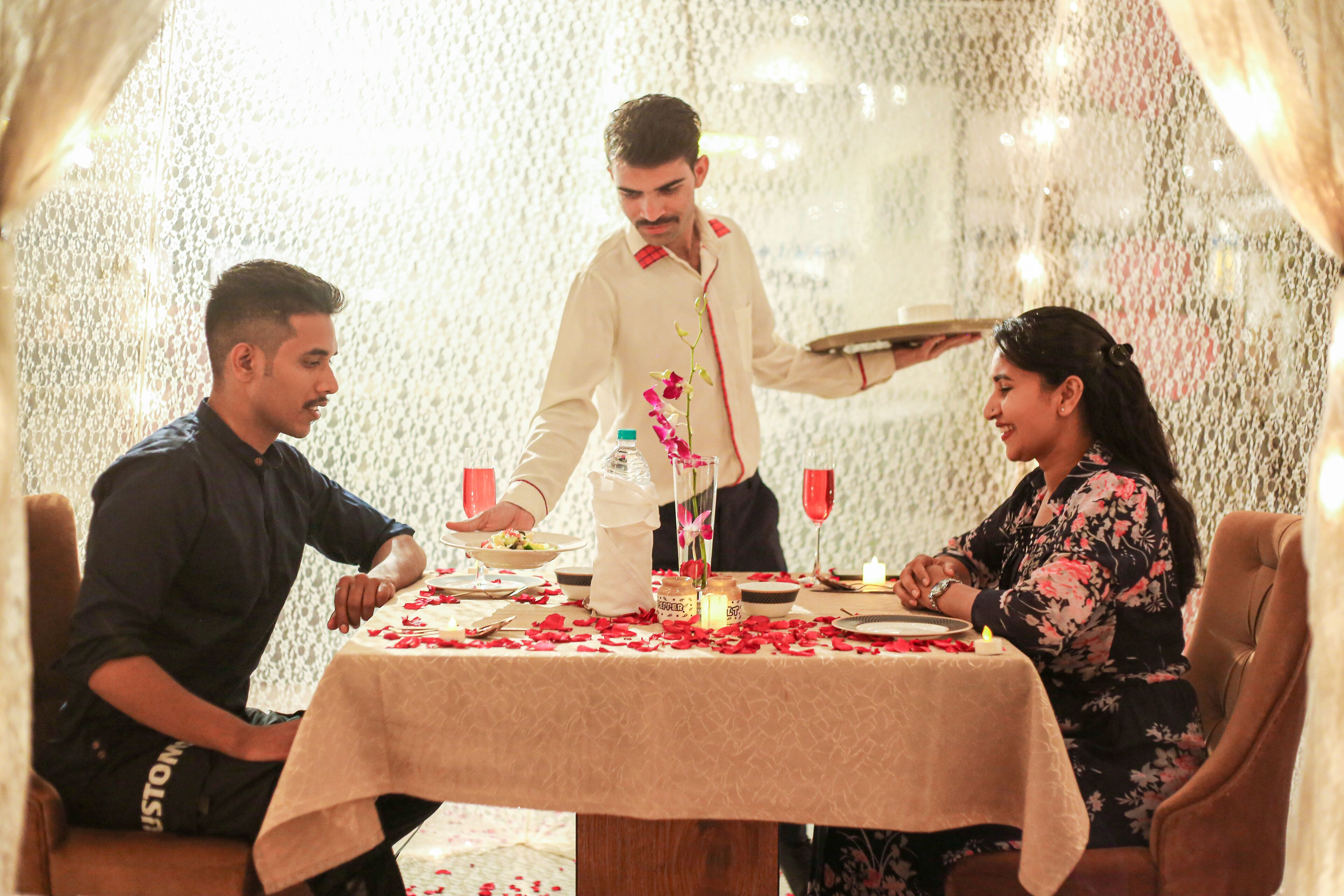 A dining table set with rose petals, candles, and a vase with flowers.