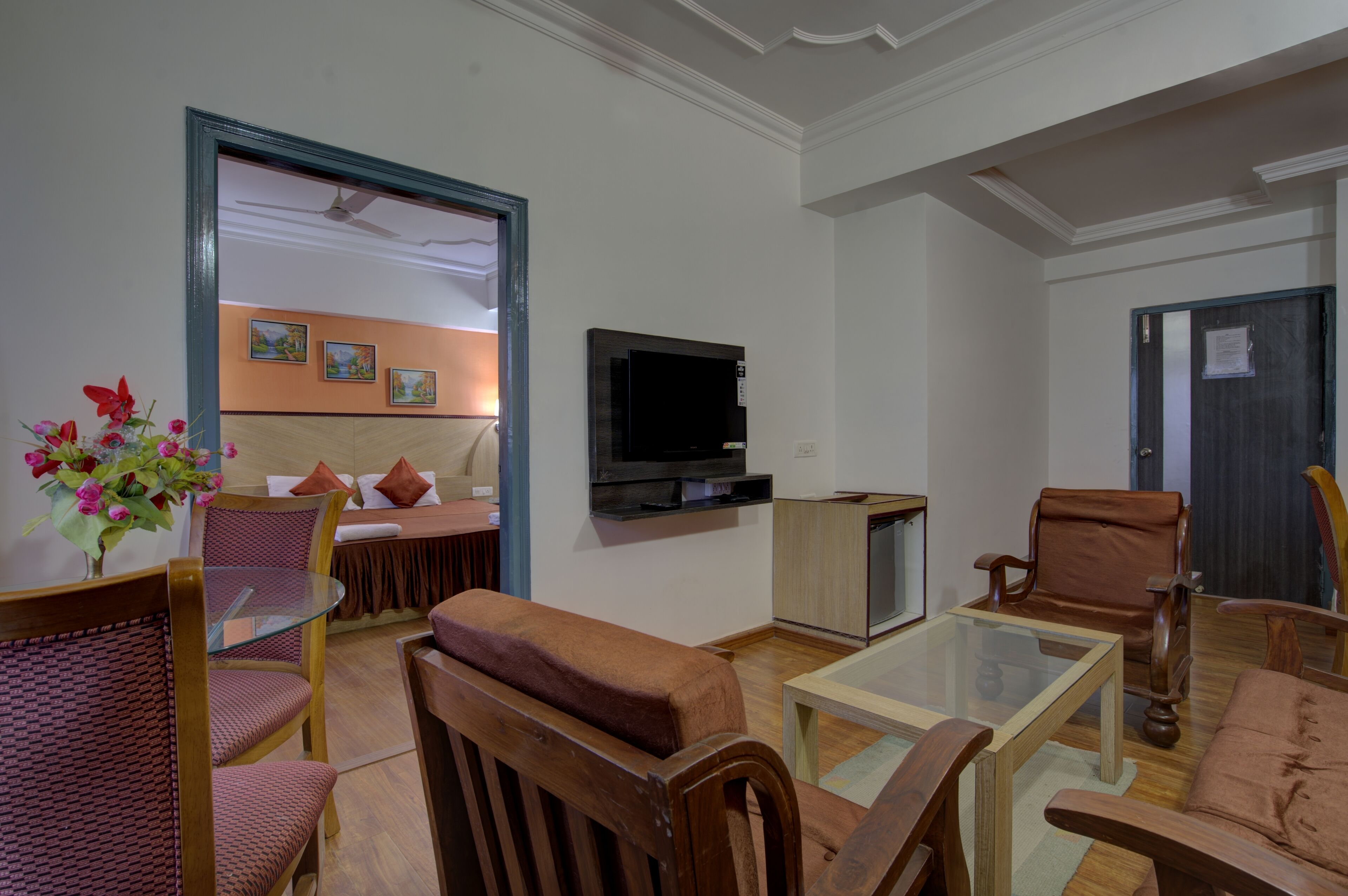A living room with a glass coffee table, wooden chairs, a sofa, a TV mounted on the wall, and a small cabinet.