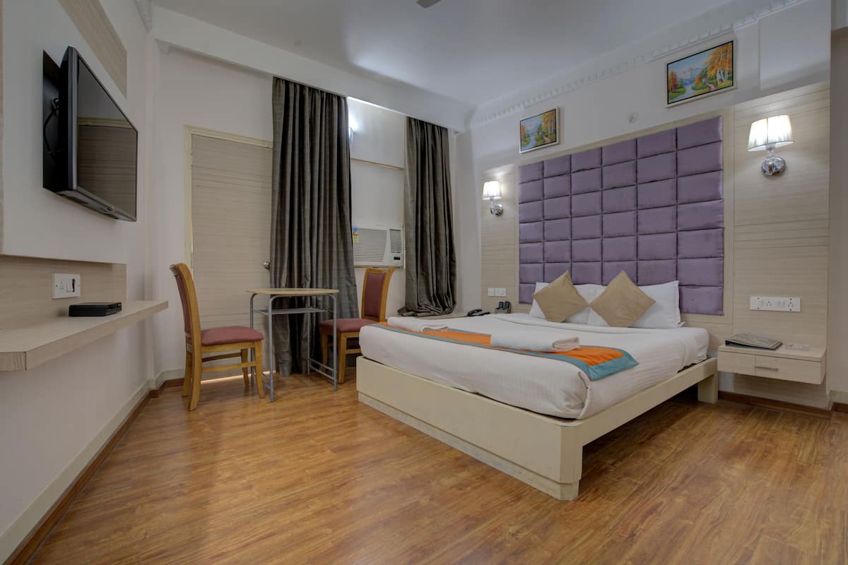 Quarto de hotel com cama, escrivaninha, cadeira, televisão e janela com cortinas.
