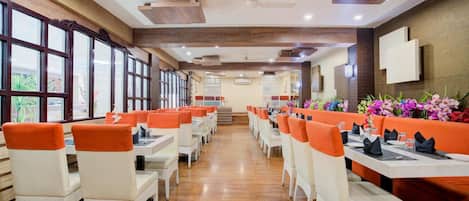 A restaurant with white wicker chairs and orange cushions, wooden floors, and large windows.