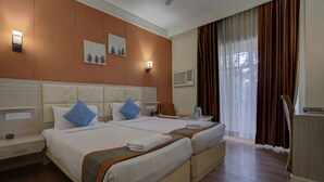 A hotel room with two beds, a nightstand, a chair, a desk, and a window with curtains. - Hotel Mahadev Palace (Deoghar)
