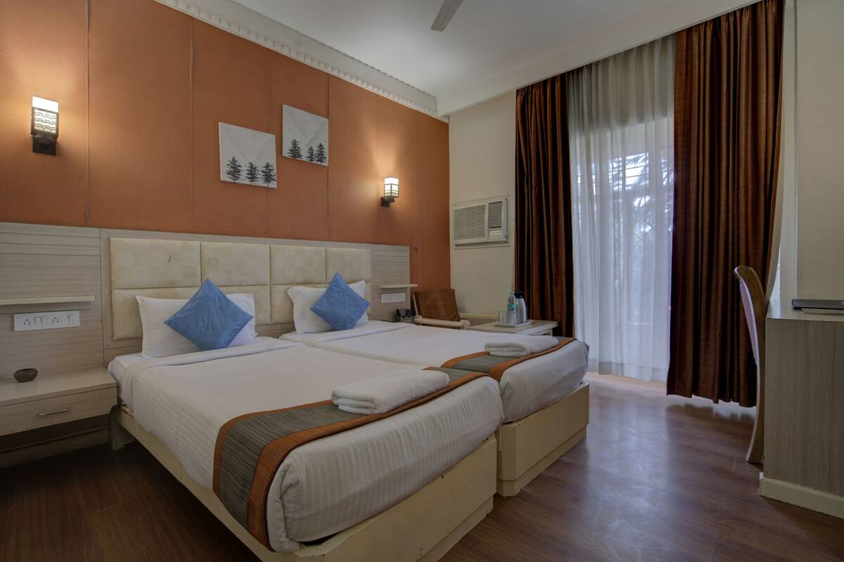 Quarto de hotel com duas camas, uma mesa de cabeceira, uma cadeira, uma escrivaninha e uma janela com cortinas.