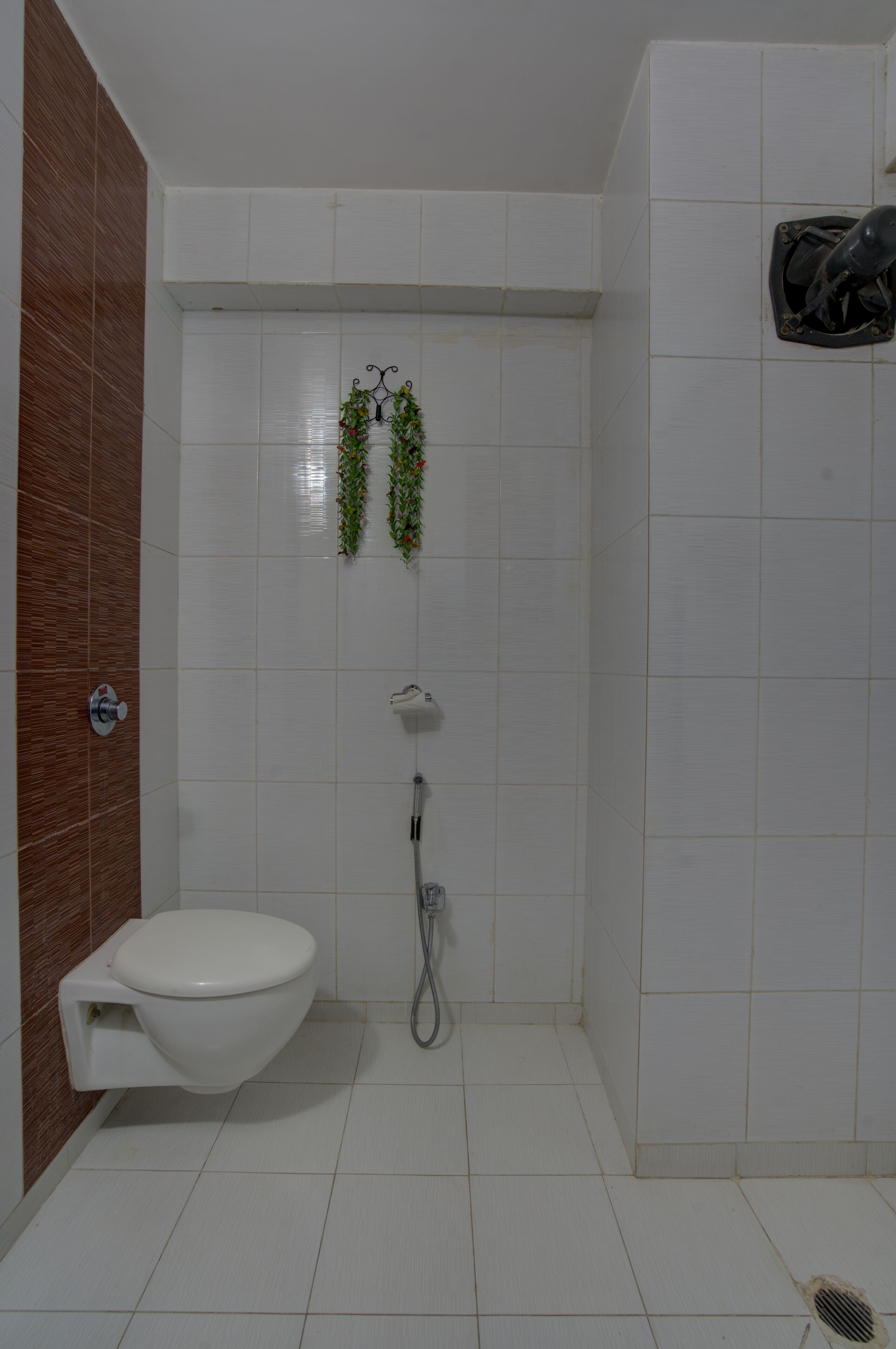 Casa de banho com azulejos brancos, sanita branca, chuveiro com chuveiro manual e toalheiro fixo com plantas.