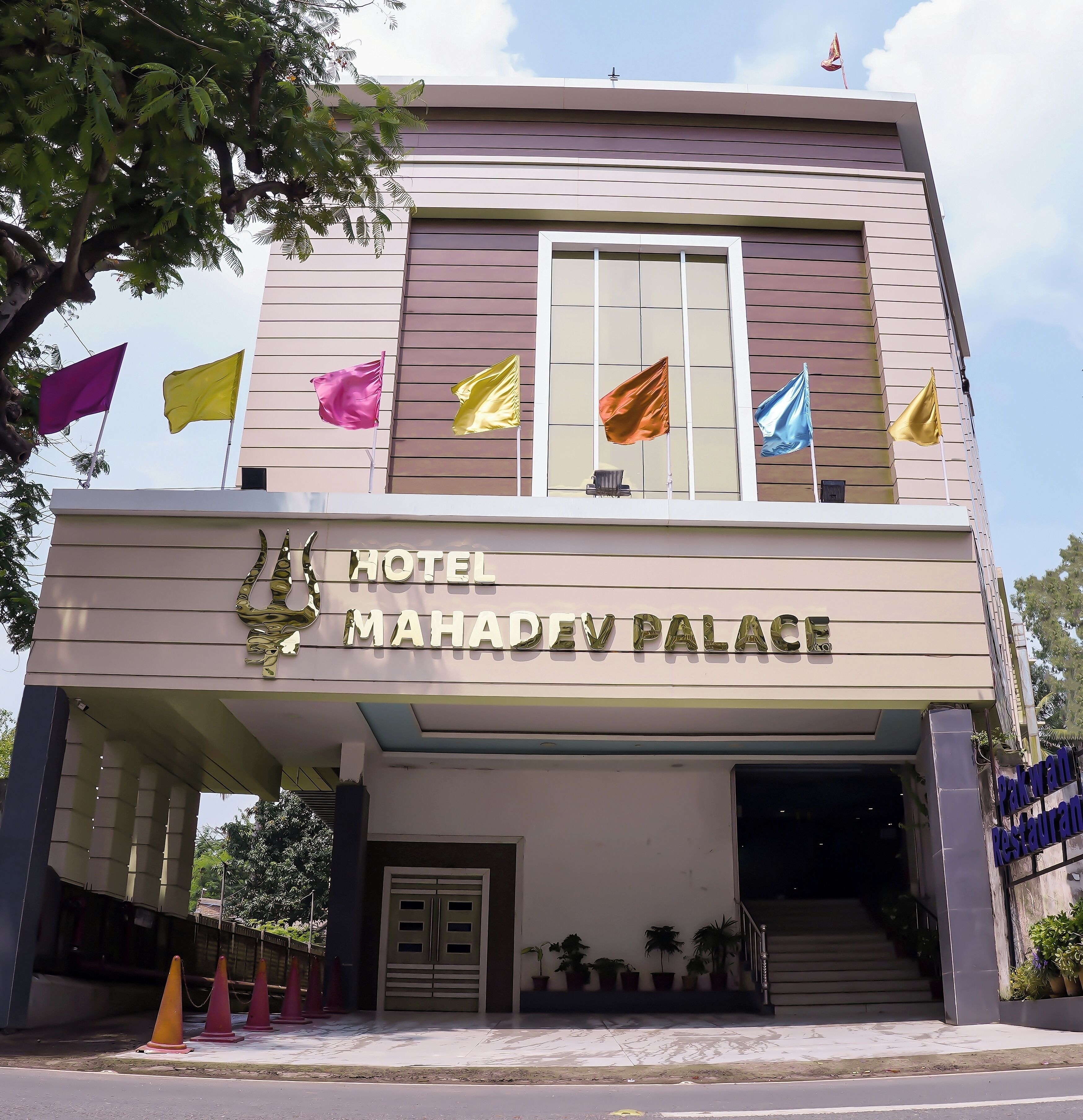 Un hotel moderno con una entrada destacada, varias banderas y un cartel que dice 'Hotel Mahadev Palace'.