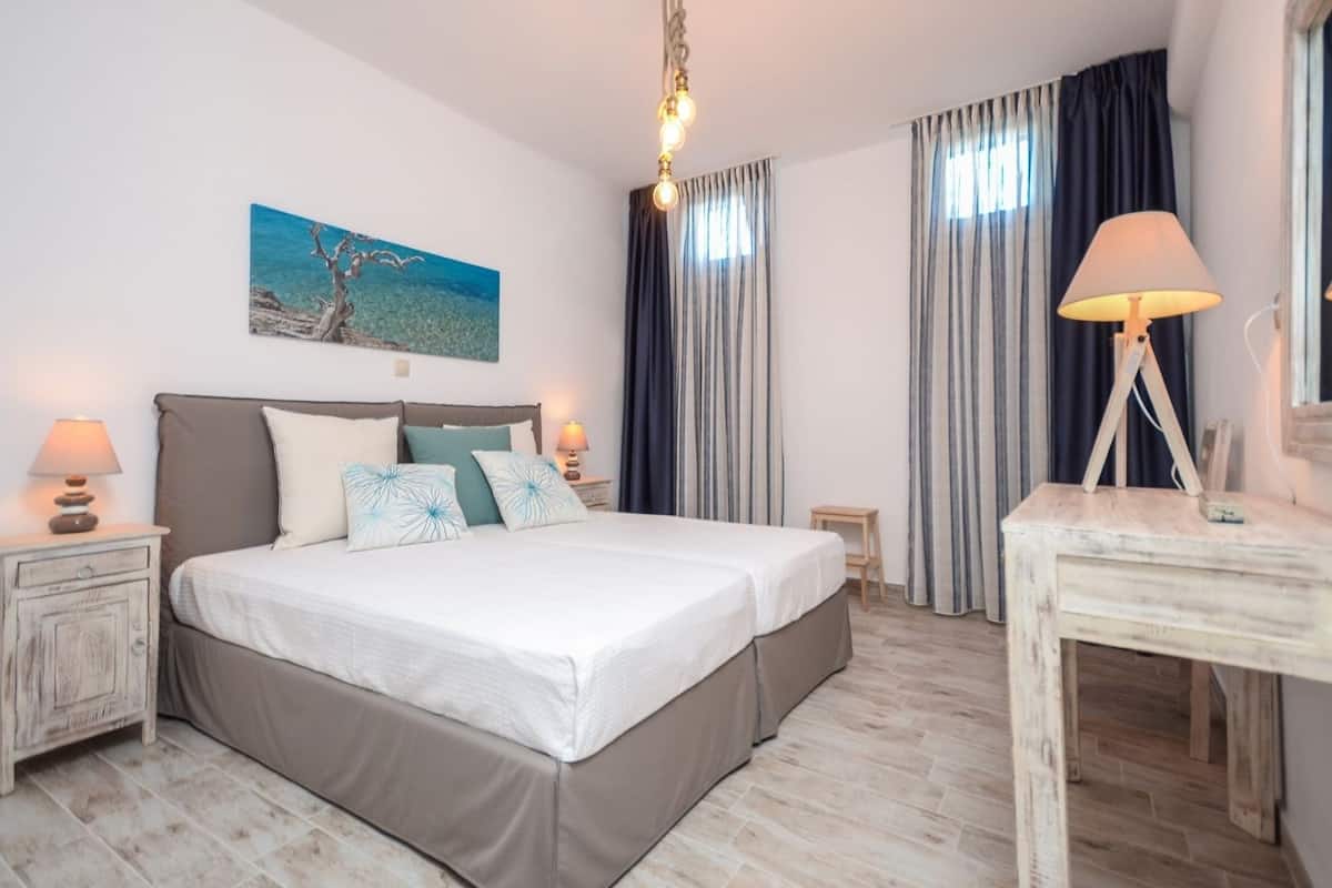 A double bed with a headboard, bedside tables with lamps, a desk with a lamp, a painting on the wall, and a view of the sea through curtains.