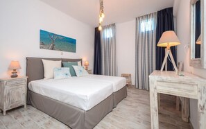 A double bed with a headboard, bedside tables with lamps, a desk with a lamp, a painting on the wall, and a view of the sea through curtains. - Aerides Villas (Naxos)