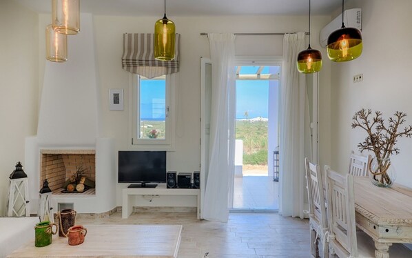A spacious living room with a fireplace, a television, and a view of the sea through an open door.