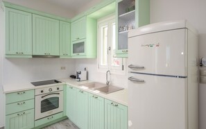 A kitchen with light green cabinets, a white refrigerator, a sink, a stove, and a window with a view of greenery.