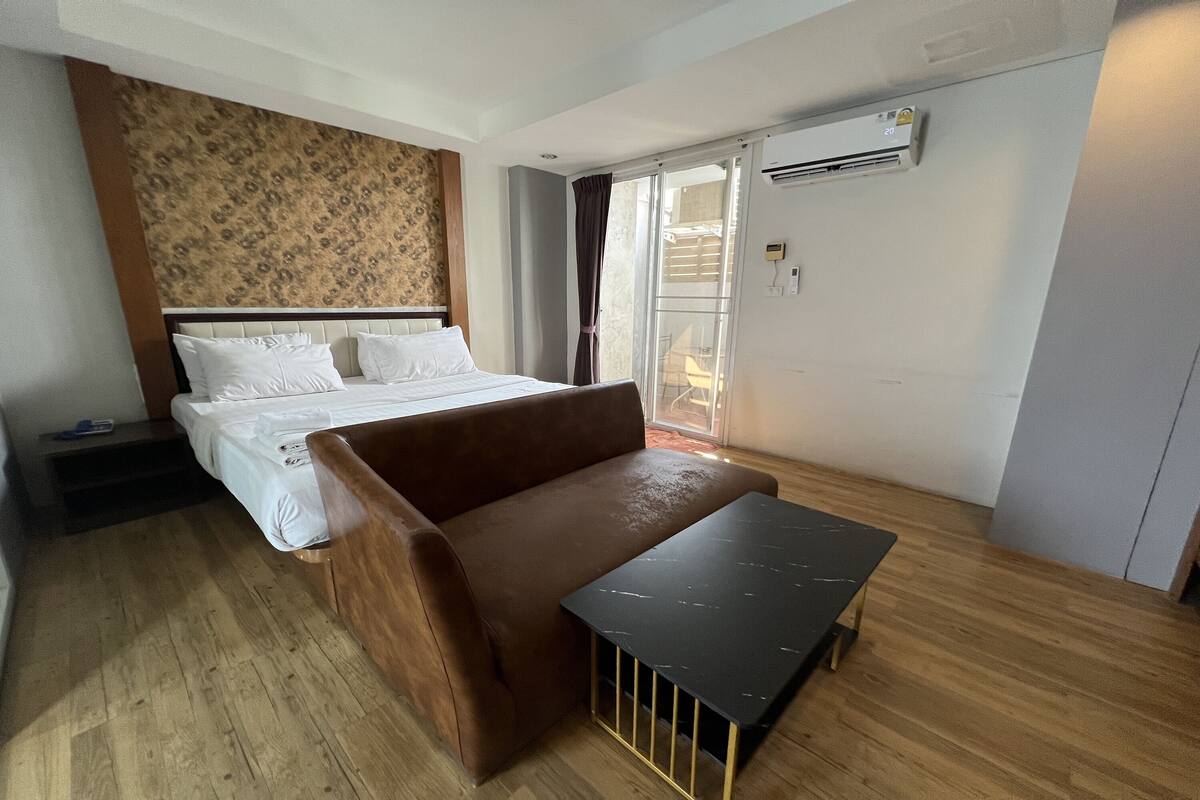 A modern hotel room with a bed, a brown sofa, a small table, and a sliding glass door leading to a balcony.