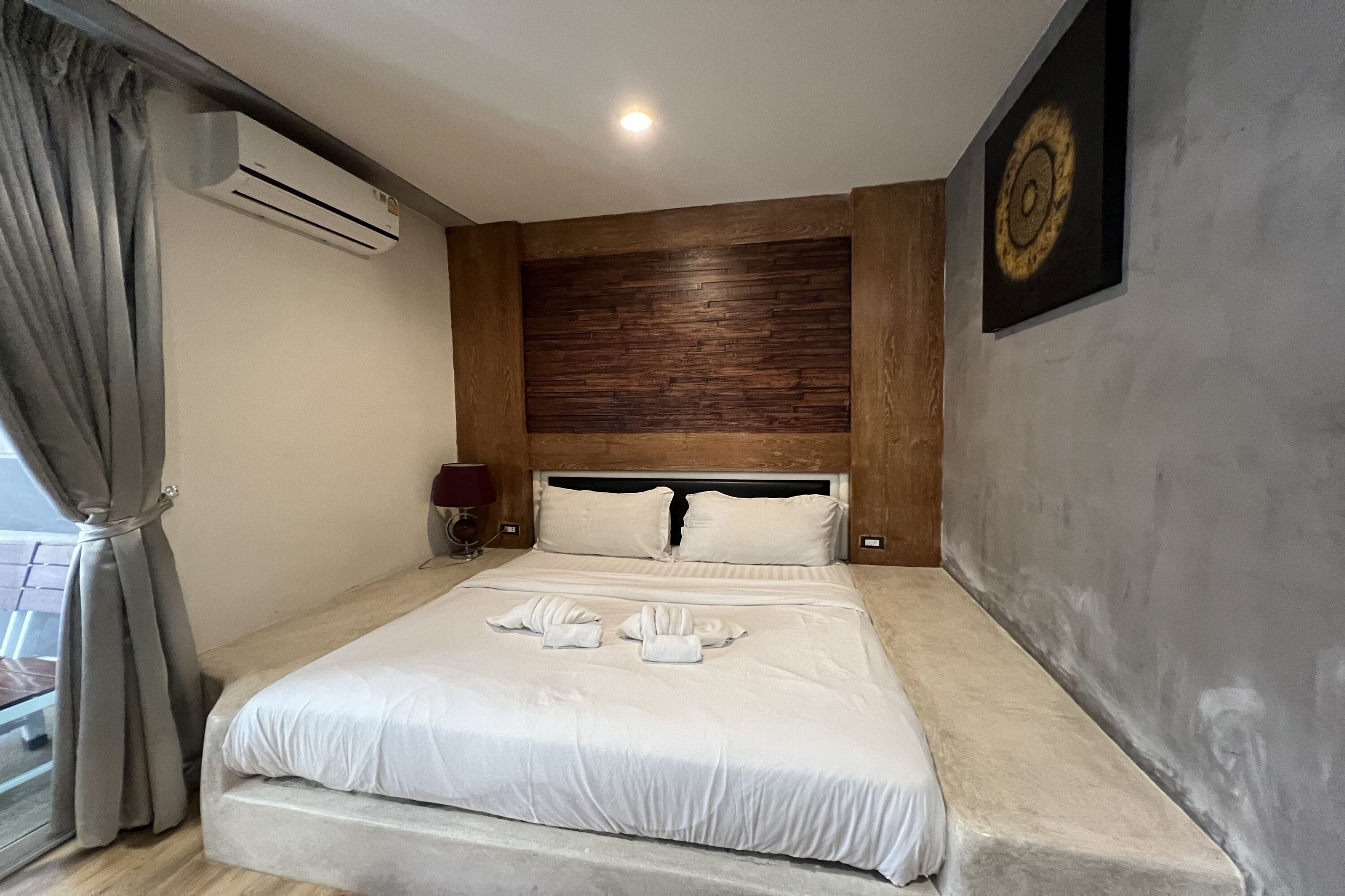 A hotel room with a large bed, a wooden headboard, a wall-mounted air conditioner, a bedside lamp, and a framed picture on the wall.